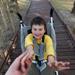 A smiling boy in a mobility stroller reaching out on a wooden boardwalk